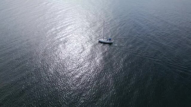 A yacht sails through the sea with its engine running and sails down in the windless weather, seen from above.