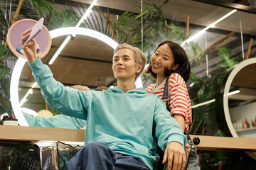 Portrait of smiling Asian man taking selfie in beauty salon with hairstylist, copy space