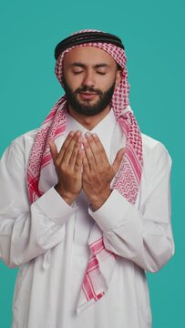 Vertical video Islamic man praying to allah in studio, having hope in religious ritual over blue background. Middle eastern person worshipping God with meditation, faithful muslim devotion.