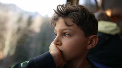 Melancholic young boy sitting by train window looking at view with hand in chin, thoughtful pensive expression of pre-teen child traveling and daydreaming - obrazy, fototapety, plakaty