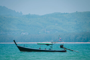 One Thai style wooden fishing boat anchored in sea close to coastline. Thai flag at boat roof damaged by strong sea winds. Boat side view