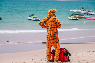 Seller dressed as orange tiger stands on a beach with big red thermal insulated bag selling ice...