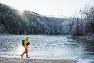 A man with a backpack walks on the shore of a frozen lake, a solo trip in winter in the mountains, trekking on ice on a pond, outdoor recreation.