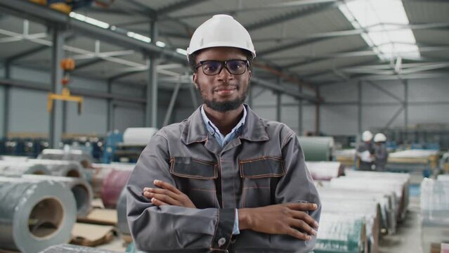 Zooming in on African American man working at industrial factory. Cheerful worker wearing safety helmet and grey uniform at workplace. Joyfully smiling while standing before camera. Occupation concept