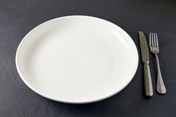 Empty white ceramic plate with cutlery on a black stone table. High angle view