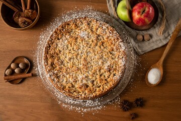 Top view of an apple crumble pie on a wooden table with cinnamon and apples in the background