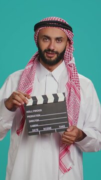Vertical video Middle eastern filmmaker holds clapboard used for scene takes on movie production, arab director using cinematography filming slate. Muslim person wearing traditional arab attire.