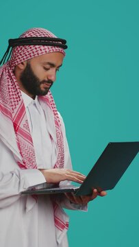 Vertical video Model using pc touchpad in studio, dressed in traditional islamic costume and kufiyah. Middle eastern person holding modern sleek laptop and scrolling websites or communicating online.