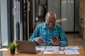 Fototapeta premium A black senior man in casual clothes uses a cell phone to find information or talk on a touch screen while working at a desk.