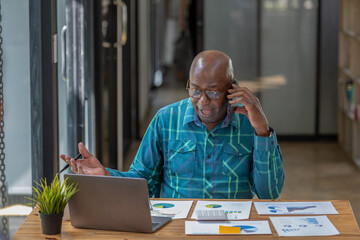 Fototapeta premium A black senior man in casual cloths using a mobile phone to discuss new season projects and business performance forecasts.
