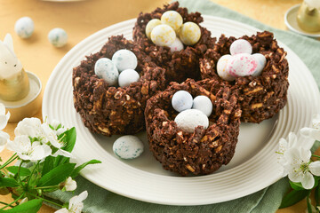 Easter chocolate nest cake with mini chocolate candy eggs with blossoming cherry or apple flowers on yellow background table. Creative recipe for Easter table with holiday decorations. Top view.