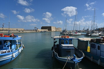 Fishing boats with white and blue wooden hulls  in Greek port of Heraklion near the city center during summer. Behind is impressive Koules Fortress.