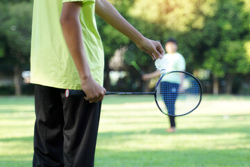 An Asian boy holds a badminton racket and a white shuttlecock while playing badminton with friends on the park lawn in the evening after returning from school. 