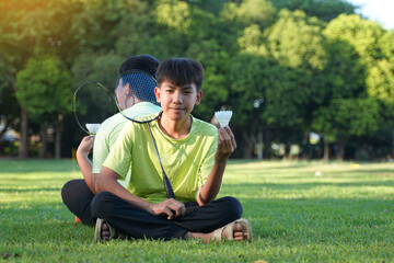 An Asian boy rests on the grass holding a badminton racket and a white shuttlecock after playing badminton with friends at the park in the evening after returning from school. 