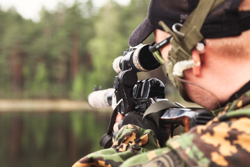 Soldier man sniper in military camouflage uniform shoots from weapon at forest with lake nature backdrop, rear view. Male border guard in country border holding weapon on war. Copy ad text space - obrazy, fototapety, plakaty