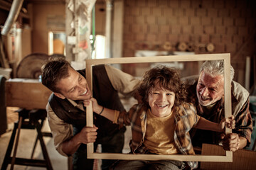 Adorable little boy posing in wood frame looking at camera with older carpenters in workshop