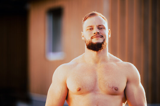 Caucasian athlete in her 30s an arrogant expression on his face stands near rural house without shirt.