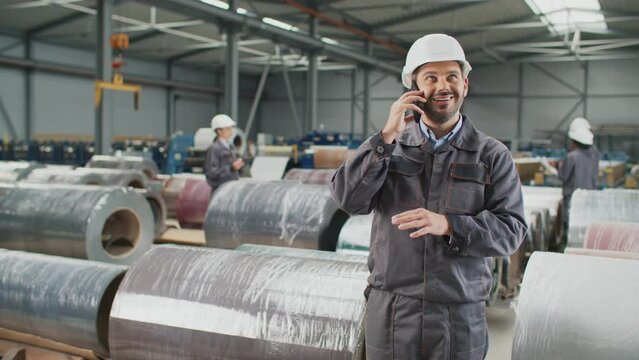 Handsome Caucasian engineer with helmet having remote conversation on his smartphone. Foreman reporting on phone to his boss about job well done in industrial production. People working in background.