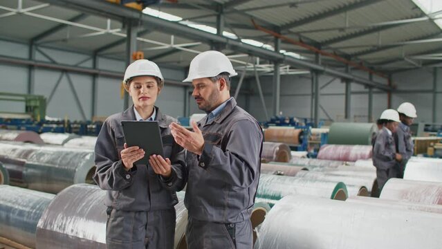 Caucasian foreman or engineer explain to his pretty assistant working process in factory. People communicating while using tablet device. In background busy workers considering industrial products.