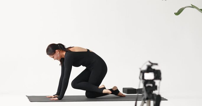 A woman blogger leading a healthy lifestyle, trains in a black one-piece sports overalls, streams for her subscribers and performs transitions with her feet while standing on all fours in a light stud