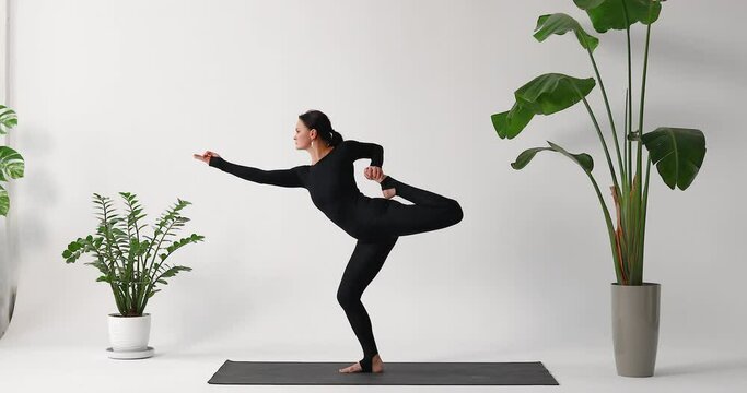 A woman practicing yoga performs a free variation of the Natarajasana exercise, dance king pose, training in a black one-piece sports jumpsuit standing on a mat in the studio on a light background