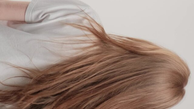 Vertical video. A red-haired girl stands with her back in the studio on a gray background and waves her hair. The concept of caring for beautiful and healthy hair.
