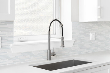 A kitchen faucet detail with a polished stainless steel faucet, a glass and stone mosaic tile backsplash, and white cabinets. - obrazy, fototapety, plakaty