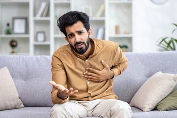 Young Hindu man holding heart sitting on sofa at home, heart attack, health problems concept, looking at camera.
