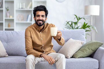 Happy sincere Arab man sitting on sofa at home holding cup in hands, drinking tea or water, concept...
