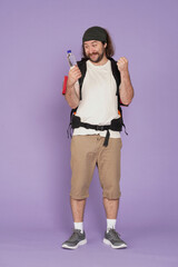 Man tourist in casual clothing with backpack holding bottle of water with happy expression. Isolated on purple background. Portrait of male traveler.