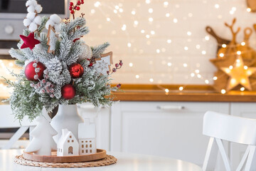 A spruce bouquet with red berries, toys, cotton on a white kitchen table against a beautiful garland. Christmas decorations in the interior. The concept of preparation for the New Year holidays 2024.