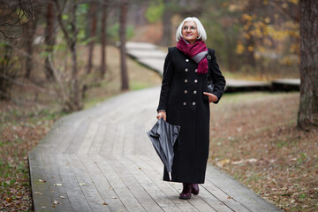 Beautiful senior woman with umbrella walks along path in forest, wears stylish trendy clothes, a warm long coat