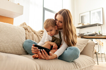 young mother and her son are sitting on the sofa and using smartphone, woman is hugging child and looking at the phone - obrazy, fototapety, plakaty