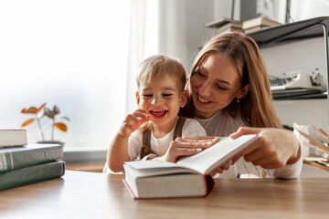 young mother with her son reads book at home, woman teaches child to read, 2 year old boy looks at book with his parent - obrazy, fototapety, plakaty