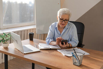 Confident stylish european middle aged senior woman using smartphone at workplace. Stylish older mature 60s gray haired lady businesswoman with cell phone in office. Boss leader using internet apps