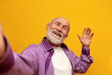 bald grandfather in purple shirt takes selfie online and greets on yellow background, old man...