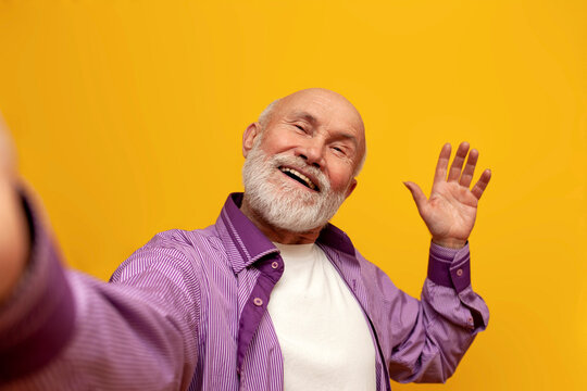 bald grandfather in purple shirt takes selfie online and greets on yellow background, old man pensioner shows palm