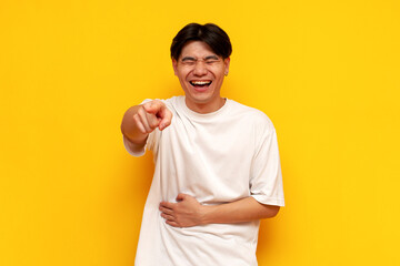 young asian guy in a white t-shirt laughs and points his hand forward on a yellow isolated background - obrazy, fototapety, plakaty