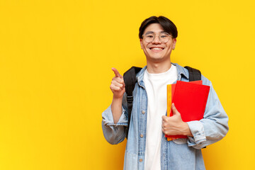 young guy asian student with backpack holds books and shows with his hand to the side on yellow...