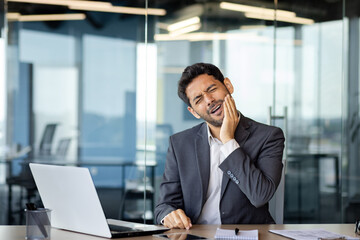 Arab businessman suffering from toothache, sitting in the office at the table in front of the laptop, holding his jaw, grimacing in pain, health concept. - obrazy, fototapety, plakaty