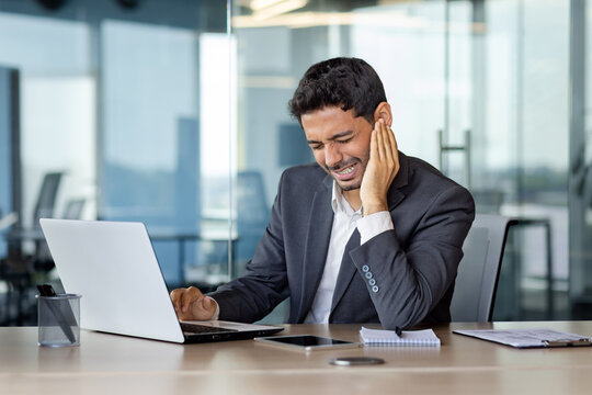 Hindu businessman suffering from ear pain, sitting in the office at the desk in front of the laptop, holding his ear, wincing in pain, health concept.