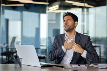 Arab businessman sitting at the table in the office holding his hand on his heart, having health...