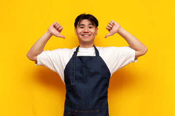 young guy asian waiter in apron shows his hands at himself on yellow isolated background