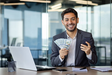 Arab man in a business suit holds a phone and a stack of dollar bills in his hands, sits at a desk in the office, rejoices at the win, smiles and looks at the camera. - obrazy, fototapety, plakaty