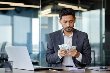 Serious Arab businessman sits in a modern office at a table in front of a laptop, counts earned...