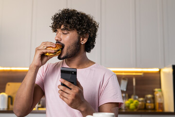 A mestizo guy stands in profile in the kitchen in subdued lighting, holds a phone and hamburger in...
