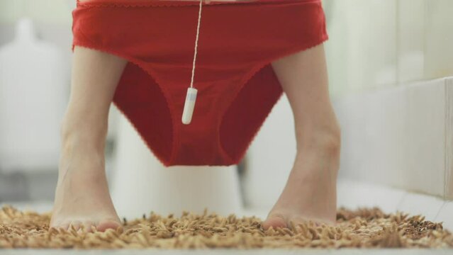 Woman sits on the toilet in a home bathroom, and shows a gynecologic tampon. Close-up of the feet and red panties. The concept of woman's health, routine hygiene and gynecology