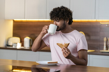 A mestizo guy stands in the kitchen in subdued lighting, holds a sandwich in hand and drinks a cup of tea or coffee with closed eyes in pleasure. A young american or african man enjoys a snack after a