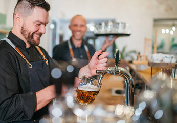 Stylish bearded barman dressed black uniform beer tapping at bar counter and waiter with tray...