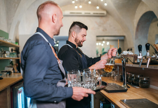 Two caucasian men friends barman beer tapping and waiter with tray dressed black uniform at bar counter. People at work, team work, men friendship and good mood concept image.